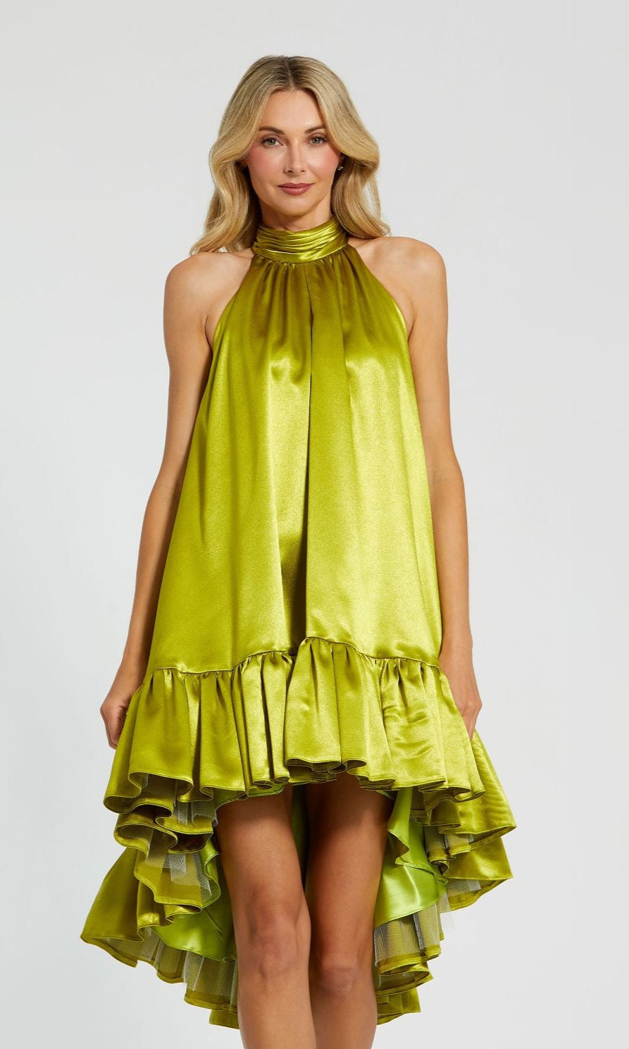 A woman models the Mac Duggal 20711 satin wedding guest dress in shiny lime green, featuring a halter neck and ruffled high-low hem, against a plain light background.